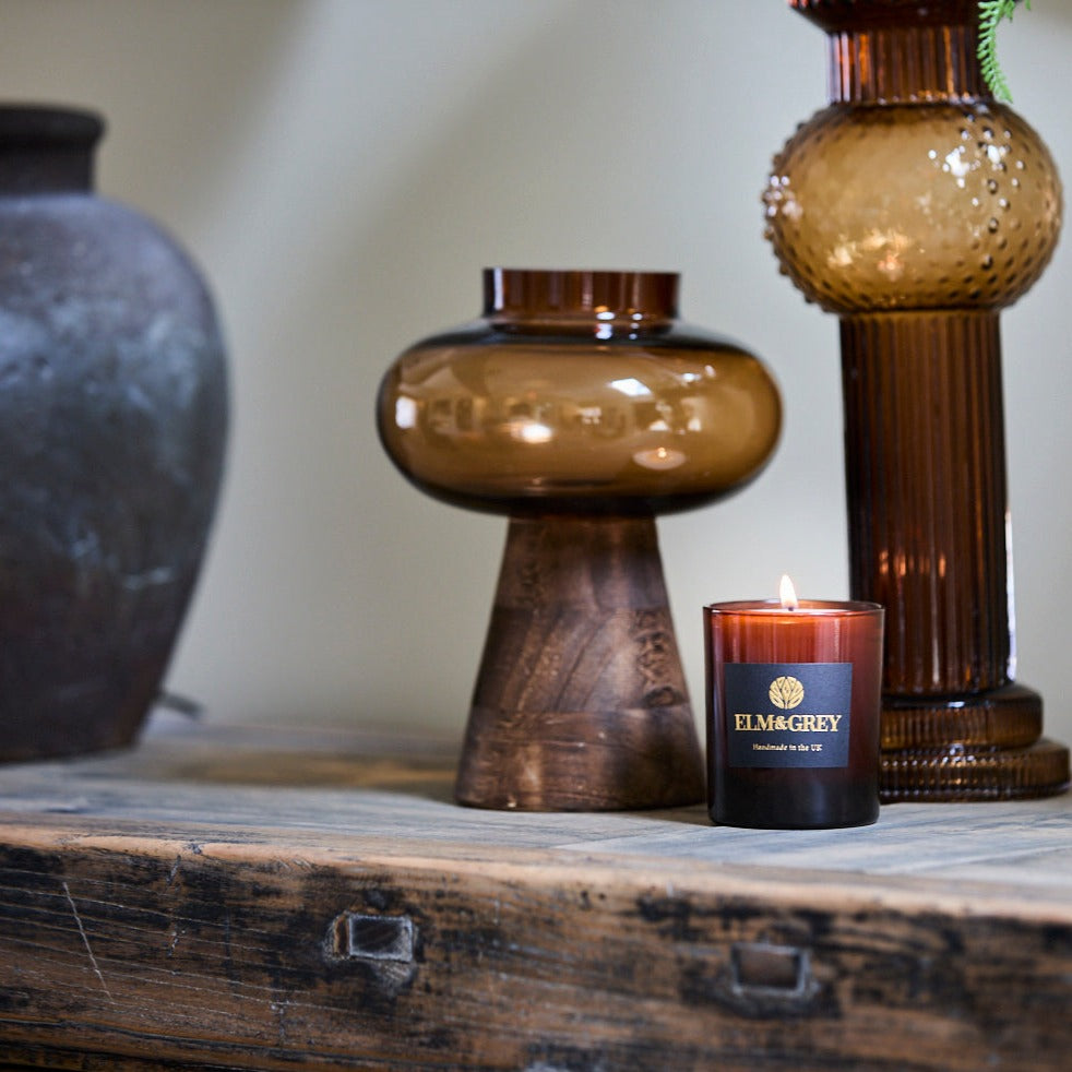 Amber glass vase on a wooden stand with a pillar candle and amber lit candle from elm and grey on a wooden sideboard